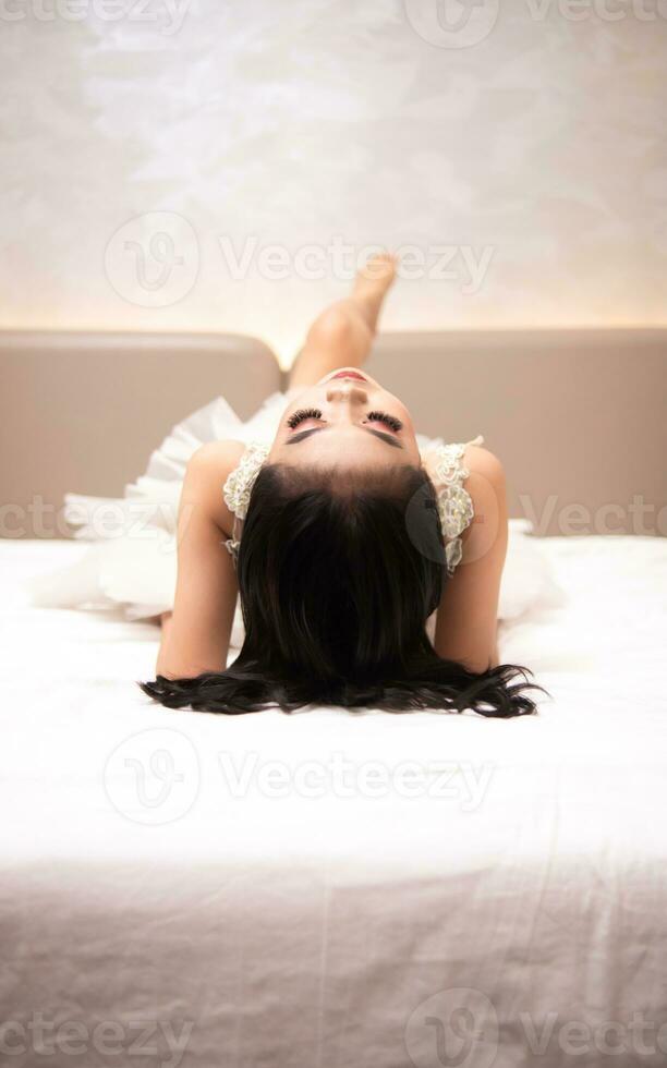 ein asiatisch Frau im ein Weiß Kleid ist Lügen auf ein Weiß Bett mit ihr schwarz Haar lose im ein Hotel foto