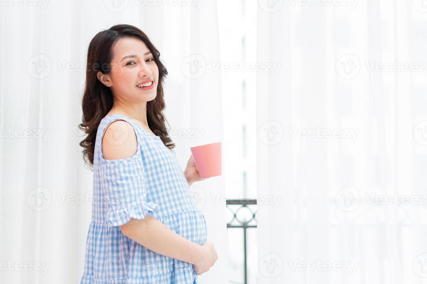 schöne frau, die ihren bauch hält und eine tasse milch hält, steht am fenster foto