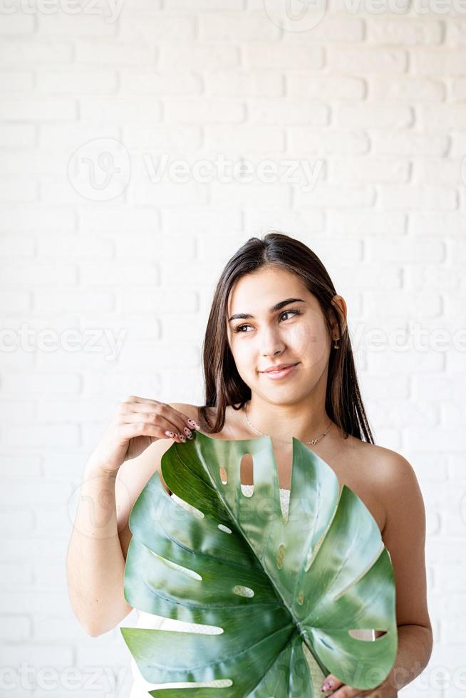 glückliche schöne Frau mit Badetüchern, die ein grünes Monstera-Blatt vor ihrem Gesicht hält foto