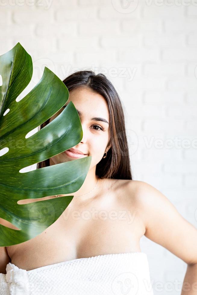 glückliche schöne Frau mit Badetüchern, die ein grünes Monstera-Blatt vor ihrem Gesicht hält foto