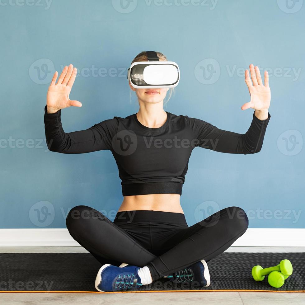 Junge blonde Frau in Sportkleidung mit Virtual-Reality-Brille, die auf der Fitnessmatte sitzt und das interaktive VR-Menü verwendet foto