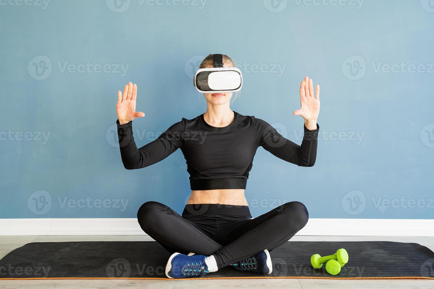 Junge blonde Frau in Sportkleidung mit Virtual-Reality-Brille, die auf der Fitnessmatte sitzt und das interaktive VR-Menü verwendet foto