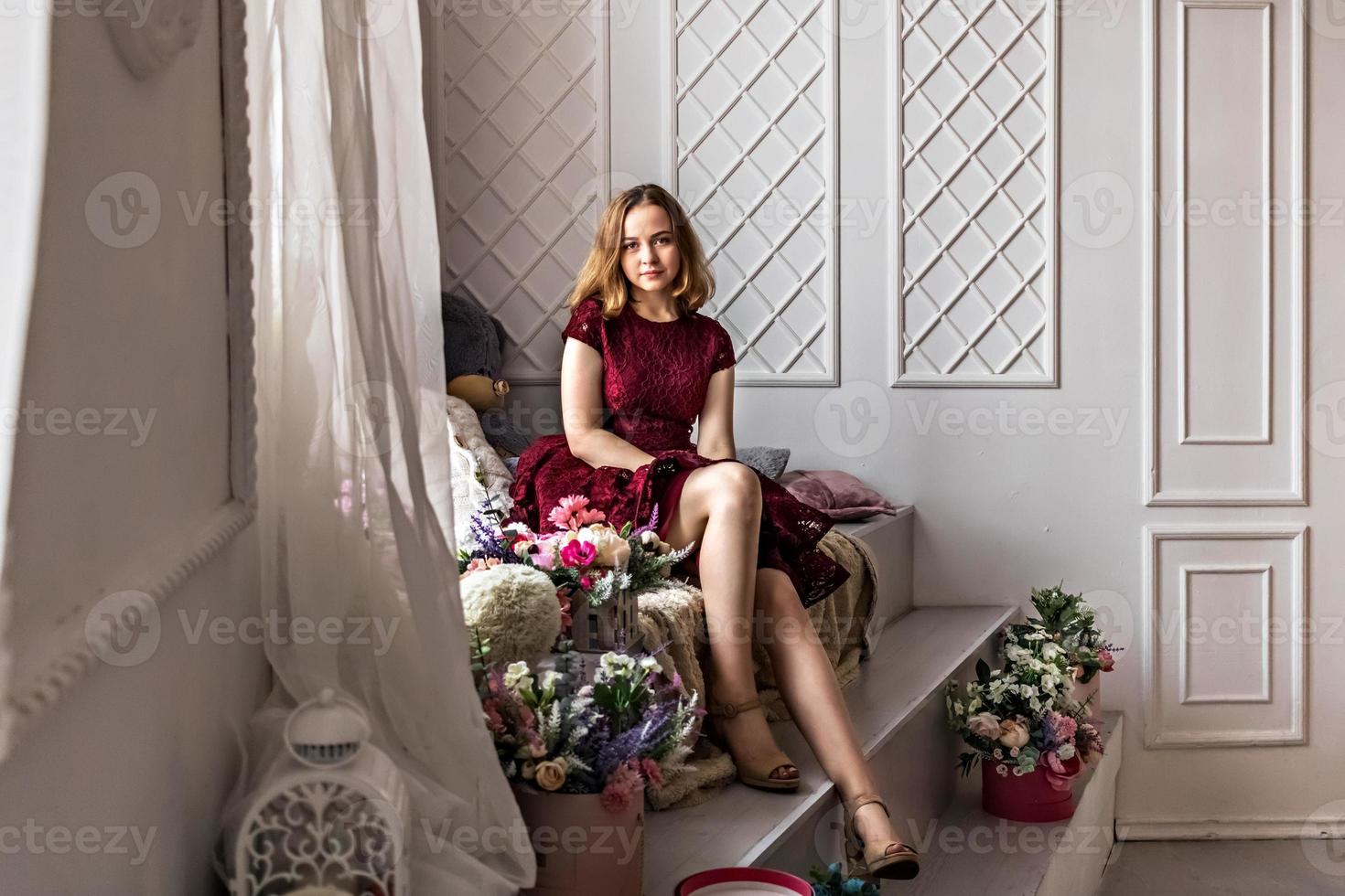 Ein süßes, stilvolles junges Mädchen in einem eleganten burgunderroten Kleid sitzt am Fenster in ihrem Zimmer. Teenager. Abschluss in Schule, College foto