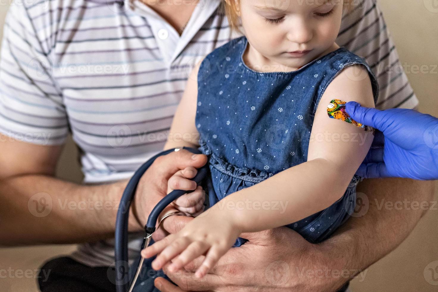 Impfung eines kleinen Mädchens in den Armen ihres Vaters in der Arztpraxis gegen das Coronavirus. lustiges klebepflaster für kinder. Impfung gegen Covid-19, Grippe, Infektionskrankheiten. foto