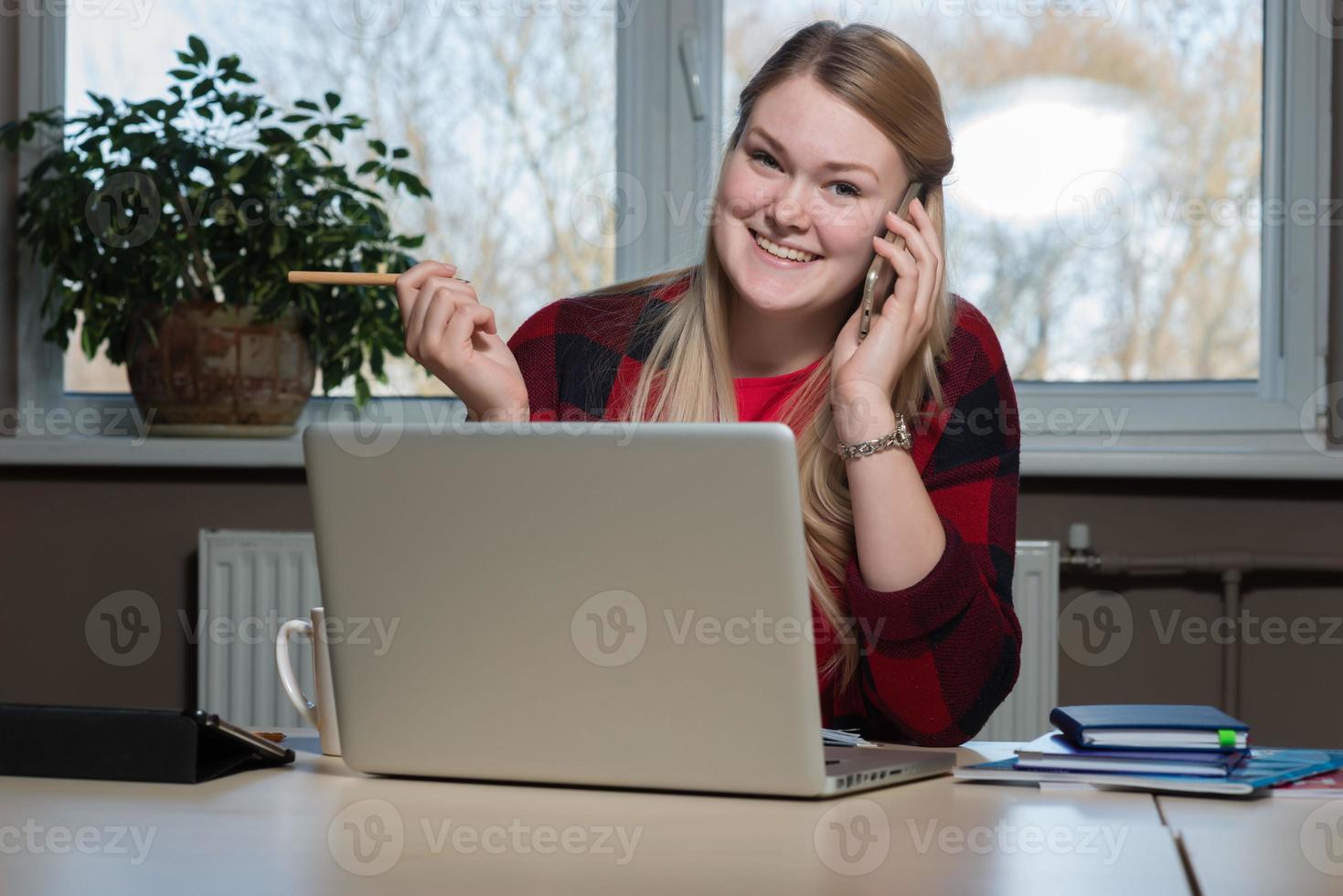 eine lächelnde blonde frau, die an einem laptop sitzt und über ein handy spricht. foto