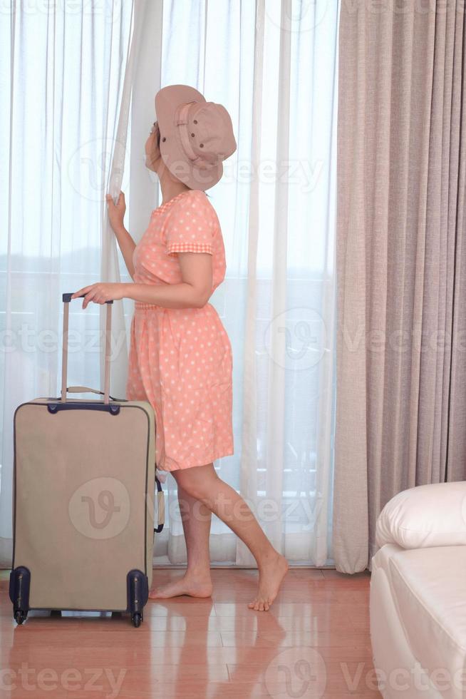 junge Reisende in rosa Kleid mit ihrem Gepäck kommt im Hotelzimmer und offenem Vorhang an, um eine Außenansicht zu genießen? foto