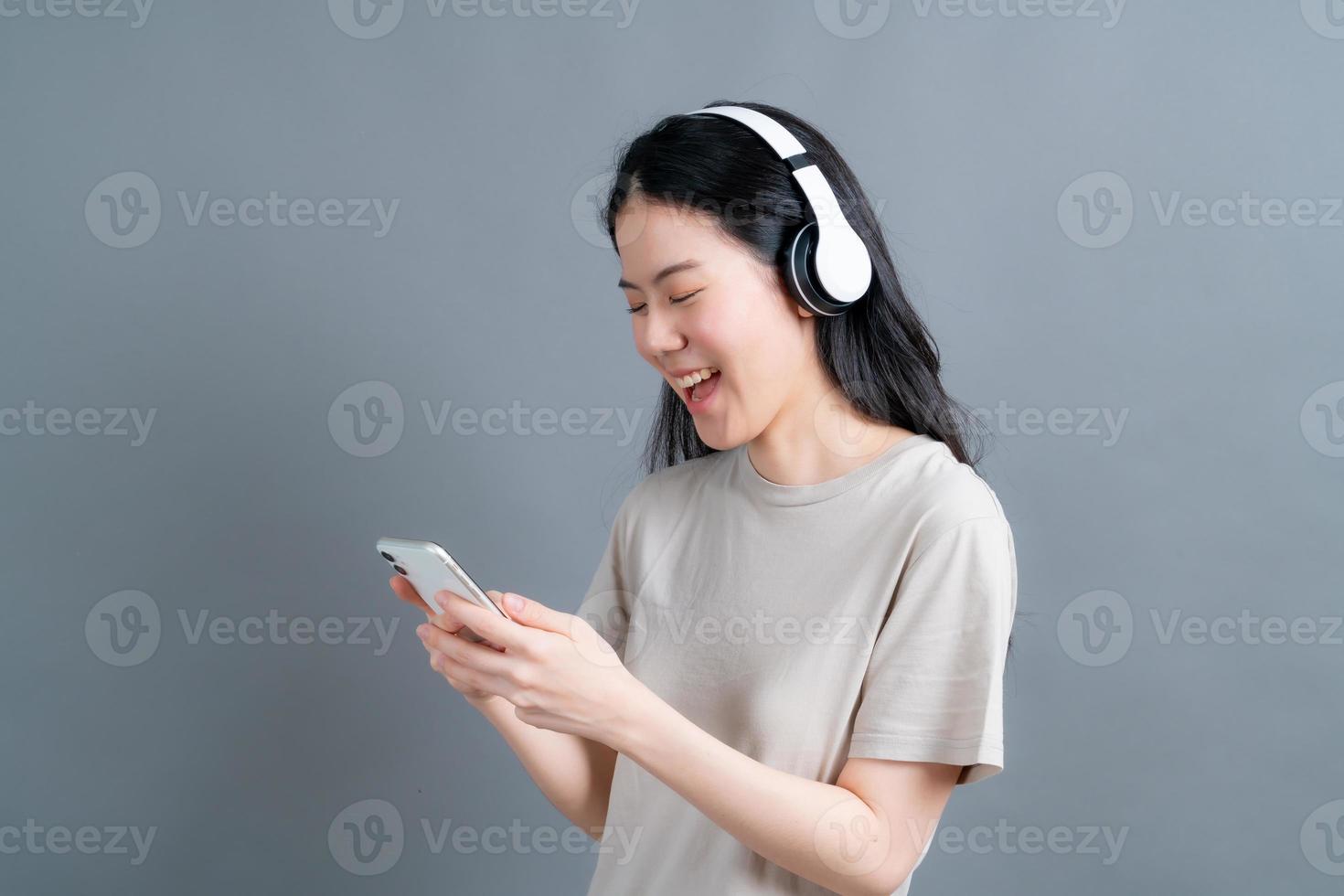 Asiatische Frau trägt einen drahtlosen Kopfhörer, der das Smartphone mit Blick auf den Telefonbildschirm mit der mobilen Player-App trägt, um Online-Musik zu hören, Fremdsprachen zu lernen, sich Videos auf grauem Hintergrund anzusehen foto