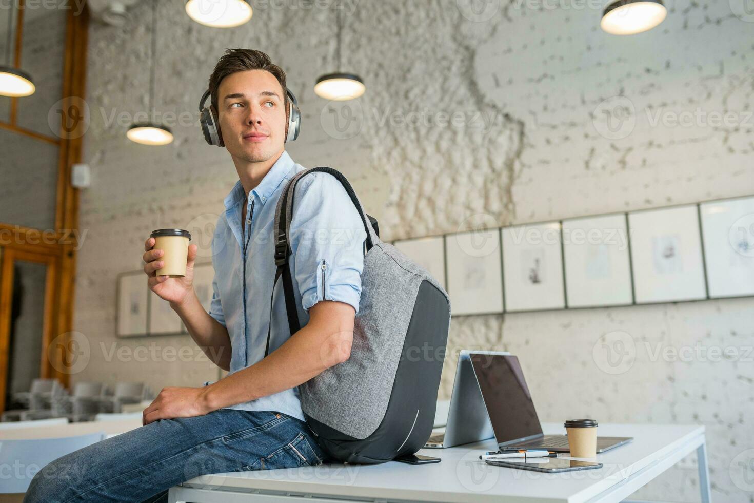 cool jung gut aussehend Mann Sitzung auf Tabelle im Kopfhörer mit Rucksack im zusammenarbeiten Büro foto