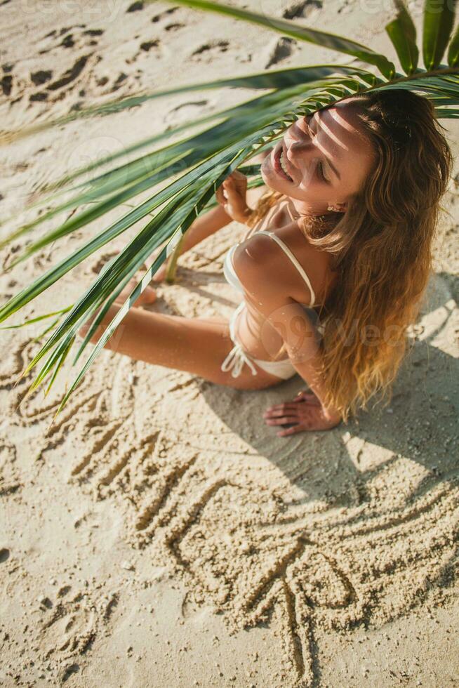 jung dünn Frau im Weiß Bikini Badebekleidung halten Blatt von Palme Baum foto