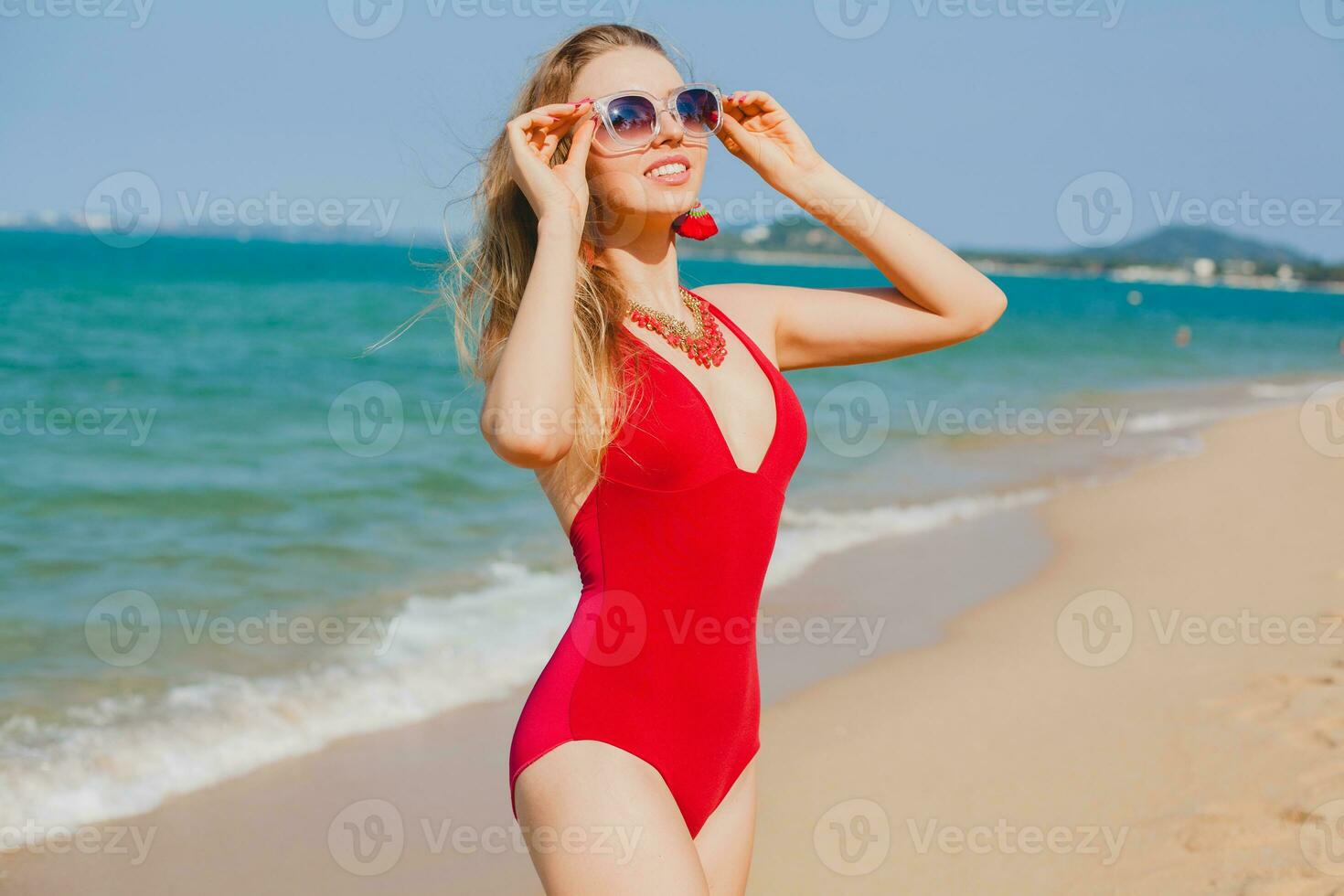 jung schön blond Frau Sonnenbaden auf Strand im rot Schwimmen Anzug, Sonnenbrille foto