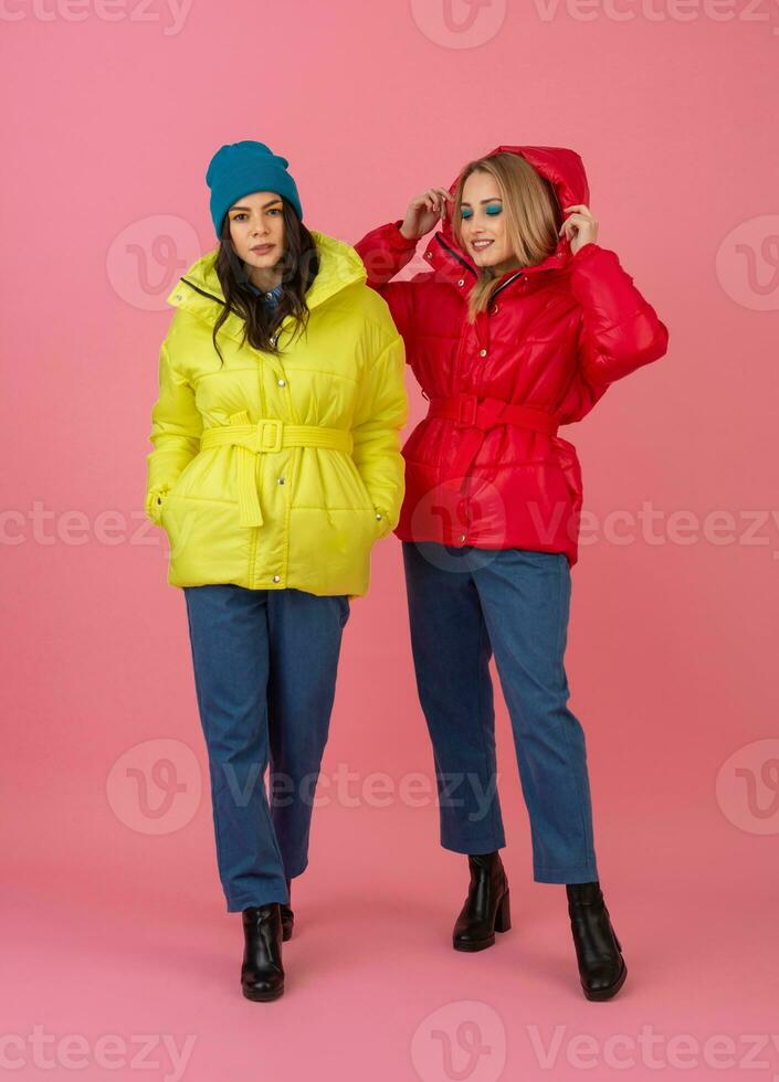 zwei aufgeregt attraktiv Mädchen freunde aktiv Frauen posieren auf Rosa Hintergrund im bunt Winter Nieder Jacke von hell rot und Gelb Farbe haben Spaß zusammen, warm Mantel Mode Trend foto