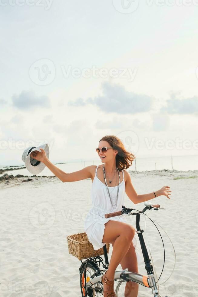 jung attraktiv lächelnd Frau im Weiß Kleid Reiten auf tropisch Strand auf Fahrrad foto