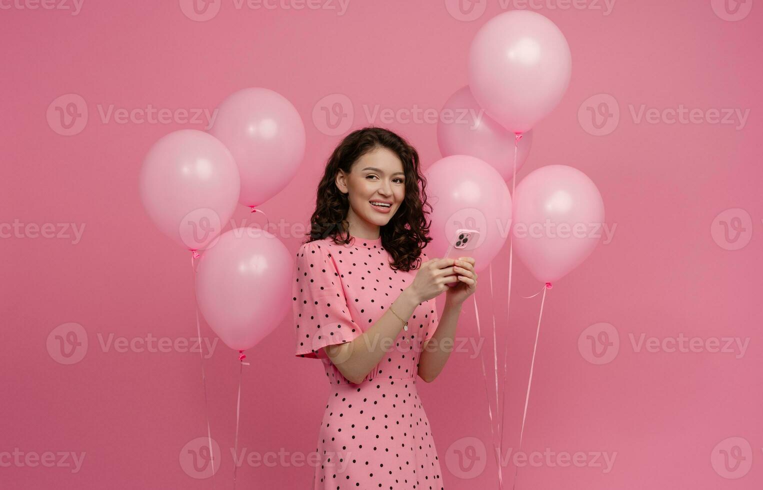 ziemlich jung Frau posieren isoliert auf Rosa Studio Hintergrund mit Rosa Luft Ballons und Smartphone foto