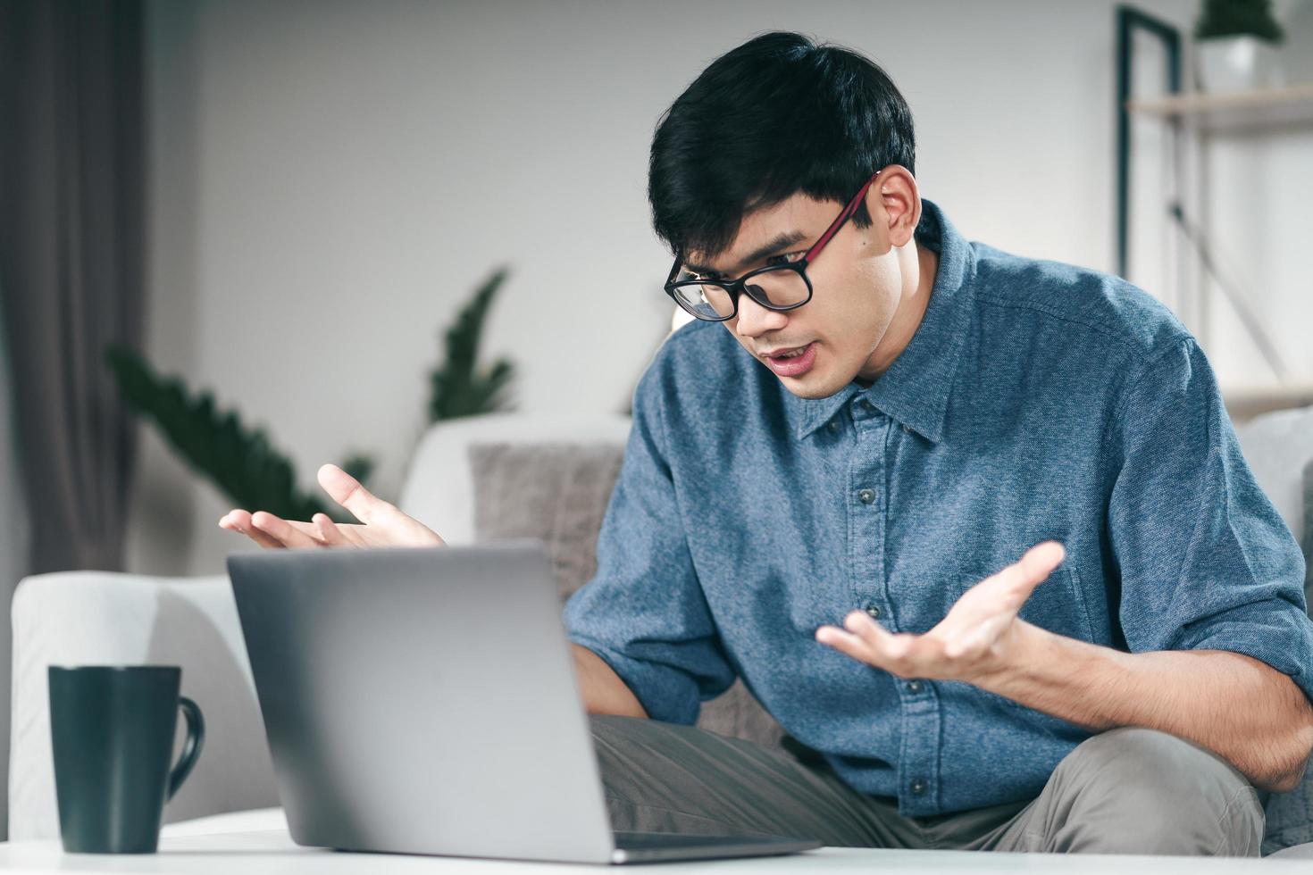 junger asiatischer Mann, der Laptop für Online-Videokonferenz verwendet, erklärt ernsthaft, nachts auf der Couch im Wohnzimmer zu sprechen. foto