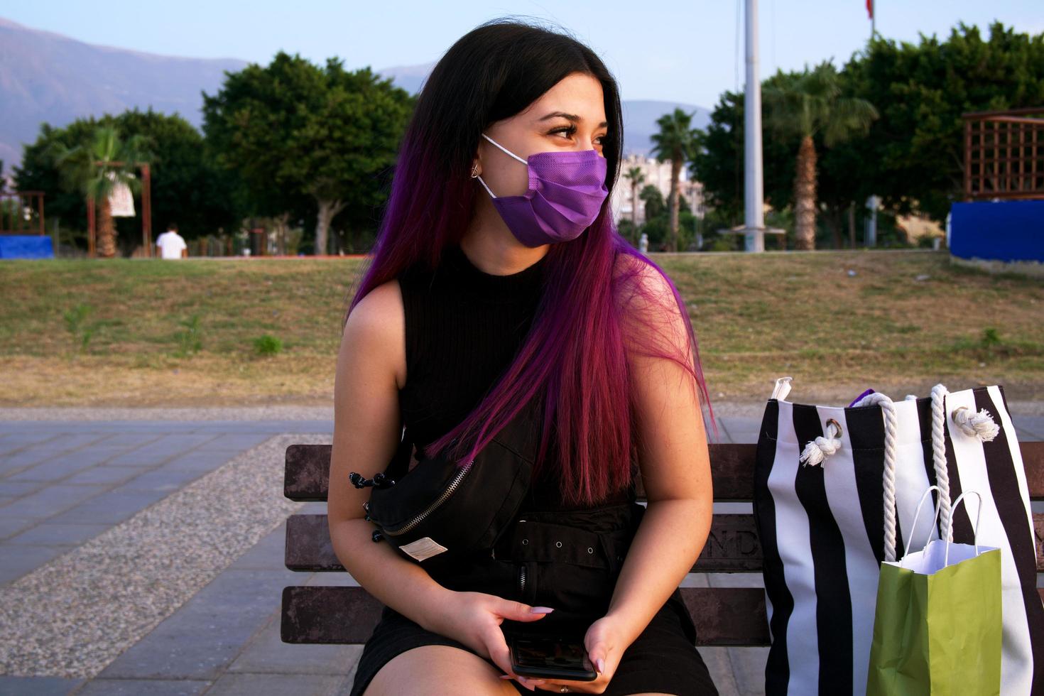 eine lilahaarige Frau mit Einkaufstüten. Shopping junge Frau ruht im Park sitzen. Sie trägt wegen Covid 19 eine lila Maske auf seinem Gesicht. foto