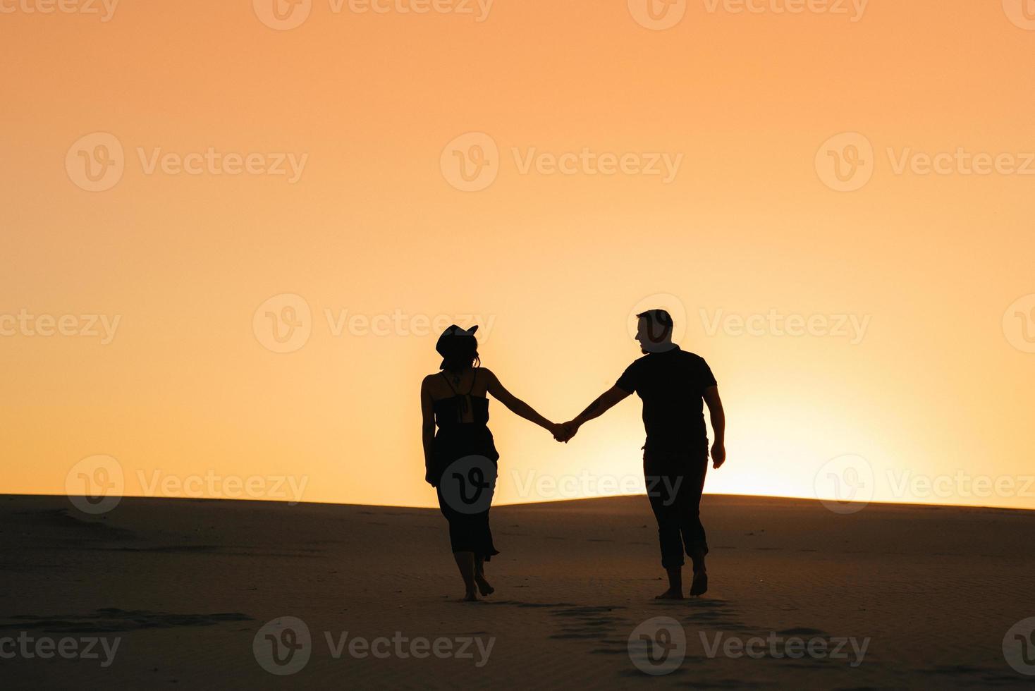 Silhouetten eines glücklichen jungen Paares auf einem Hintergrund des orangefarbenen Sonnenuntergangs in der Sandwüste foto