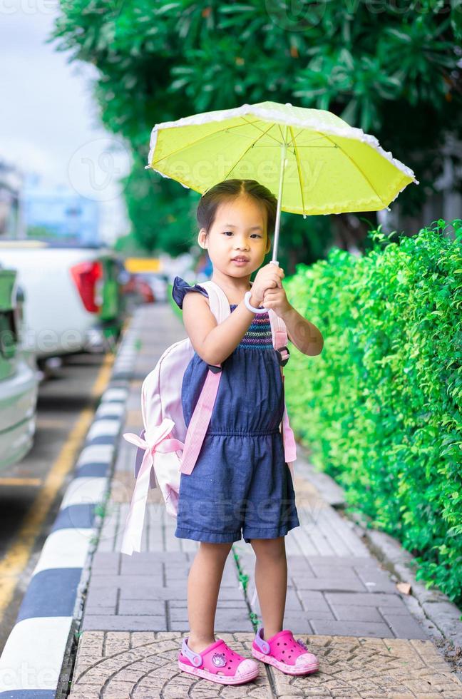 kleines Mädchen mit Regenschirm und Rucksack, das im Park am Regentag zurück zur Schule geht? foto