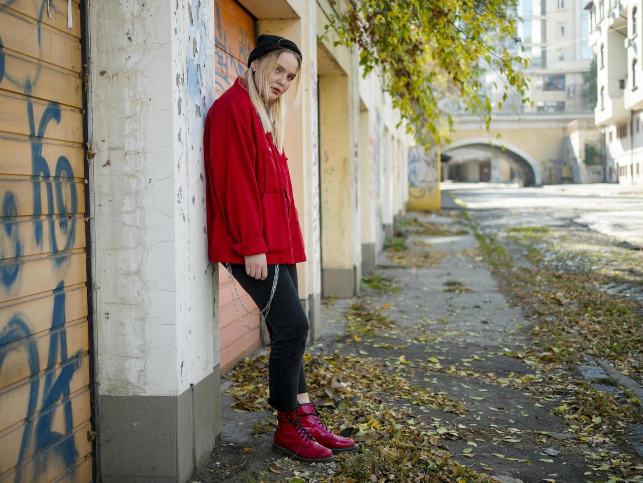 süßes Mädchen in einer roten Strickjacke und roten Stiefeln, die in der Nähe einer Wand in vollem Wachstum im Freien stehen foto