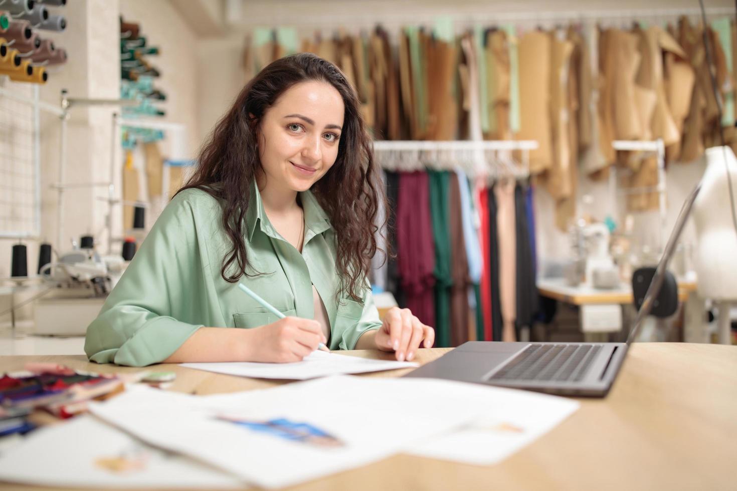 Elegante Designerin, die am Laptop arbeitet und am Schreibtisch im Studio skizziert. junge Modearbeiterin in der Werkstatt foto