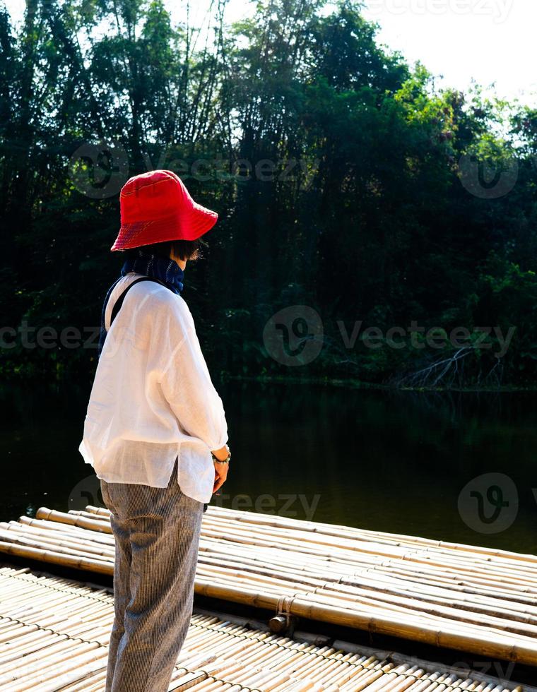 Rückansicht der einsamen Frau in Rot hatte auf dem Bambusboden gesessen und den grünen Wald und den Fluss betrachtet foto