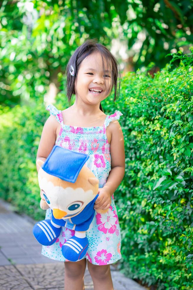 Porträt eines glücklichen asiatischen kleinen Mädchens mit Puppe, das auf einem Fußweg im Park steht foto