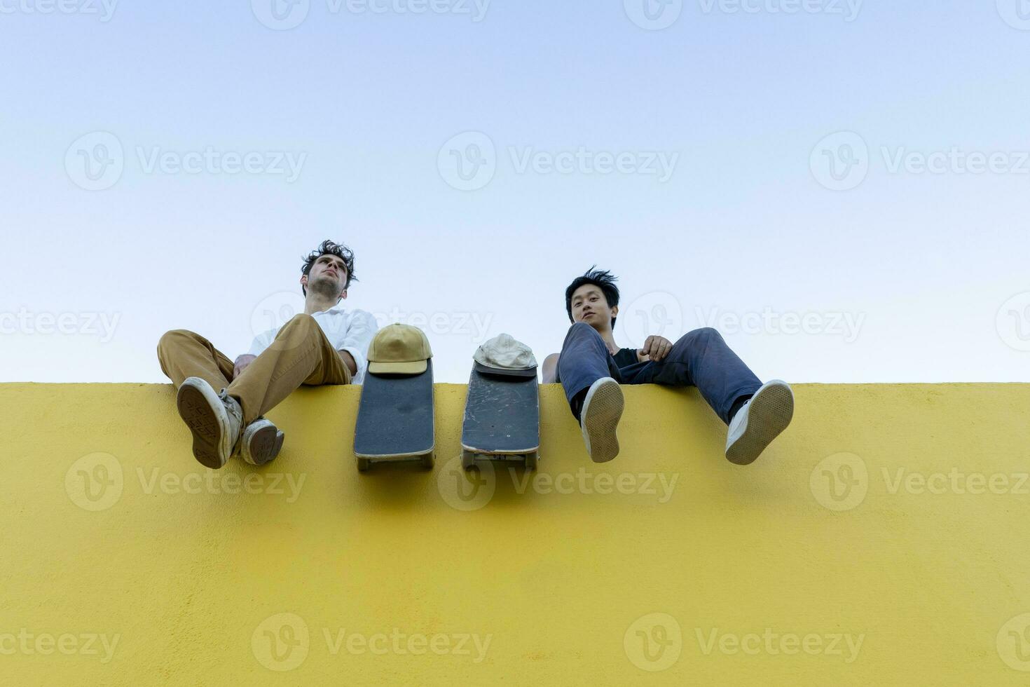 zwei jung Männer mit Skateboards Sitzung auf ein hoch Gelb Mauer foto