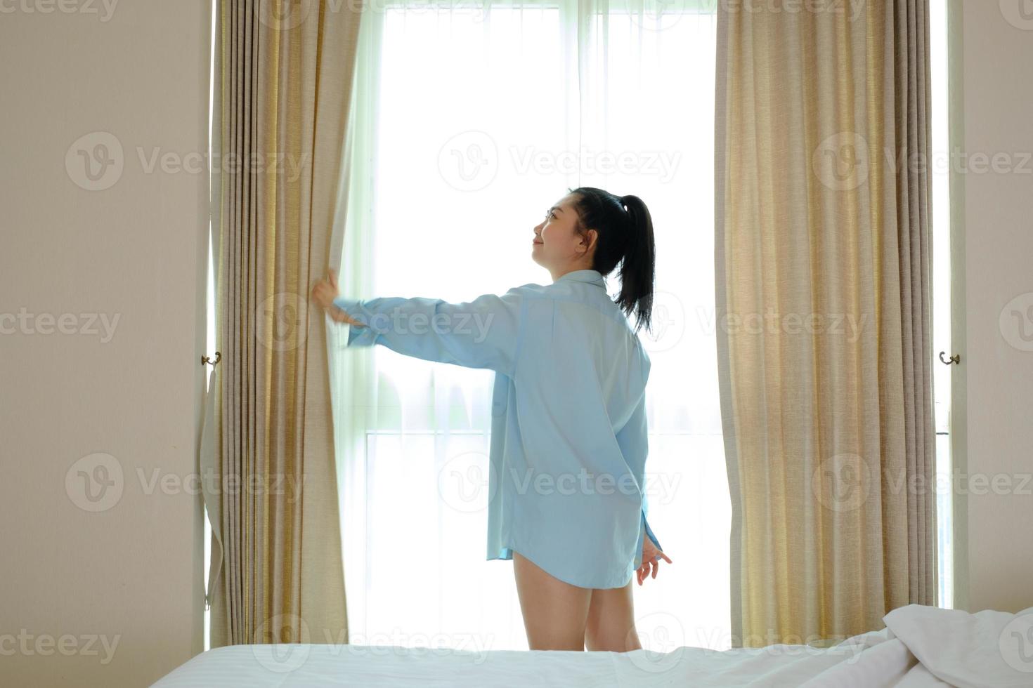 junge Frau, die morgens nach dem Aufwachen die Vorhänge im Schlafzimmer öffnet foto