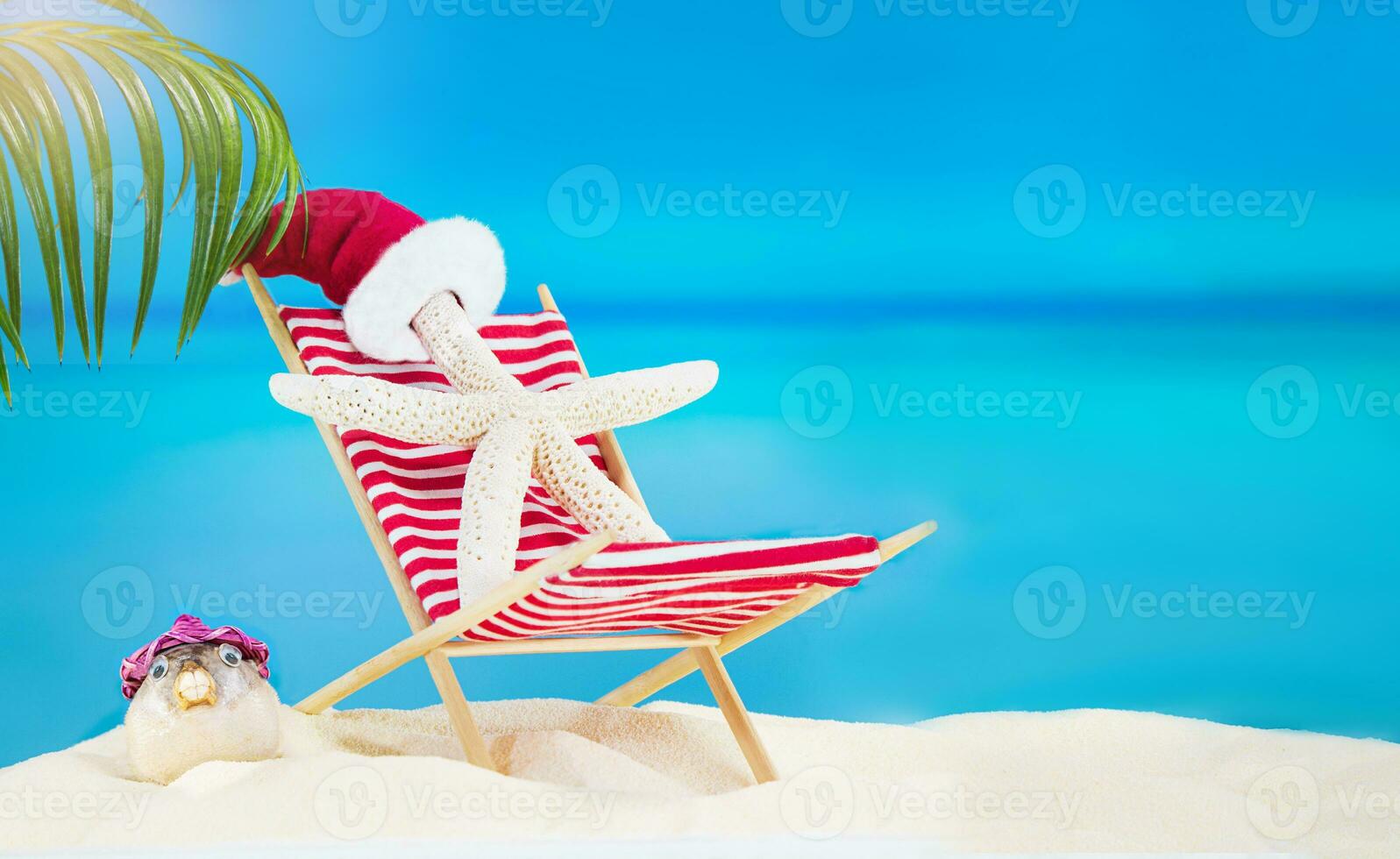 Weiß Seestern mit Weihnachtsmann Hut auf gestreift rot Sonne Liege mit Puffer Fisch auf sandig Strand mit Palme Baum hinter Meer. Weihnachten, Neu Jahr, Sommer, reisen, Urlaub, Urlaub im heiß Länder. Kopieren Raum foto