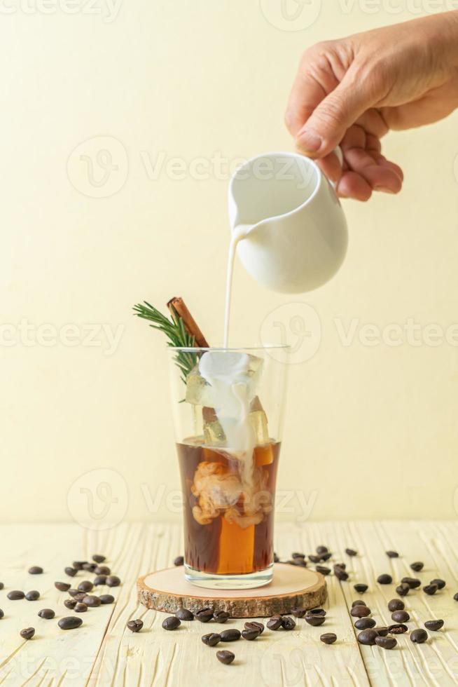 Gießen von Milch in schwarzes Kaffeeglas mit Eiswürfel, Zimt und Rosmarin auf Holzhintergrund foto