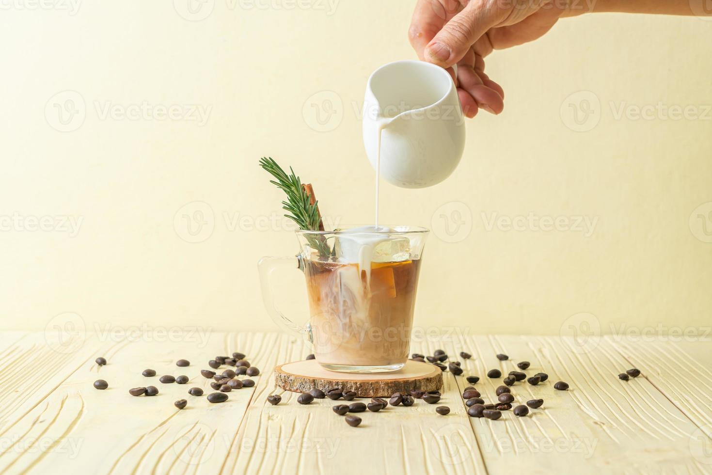 Gießen von Milch in schwarzes Kaffeeglas mit Eiswürfel, Zimt und Rosmarin auf Holzhintergrund foto