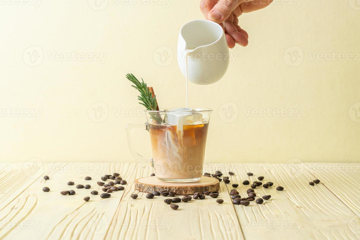 Gießen von Milch in schwarzes Kaffeeglas mit Eiswürfel, Zimt und Rosmarin auf Holzhintergrund foto