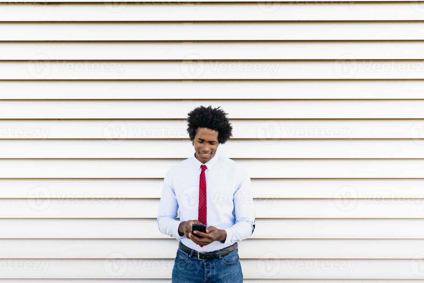 schwarzer Geschäftsmann mit einem Smartphone mit weißem Hintergrund mit Jalousien foto