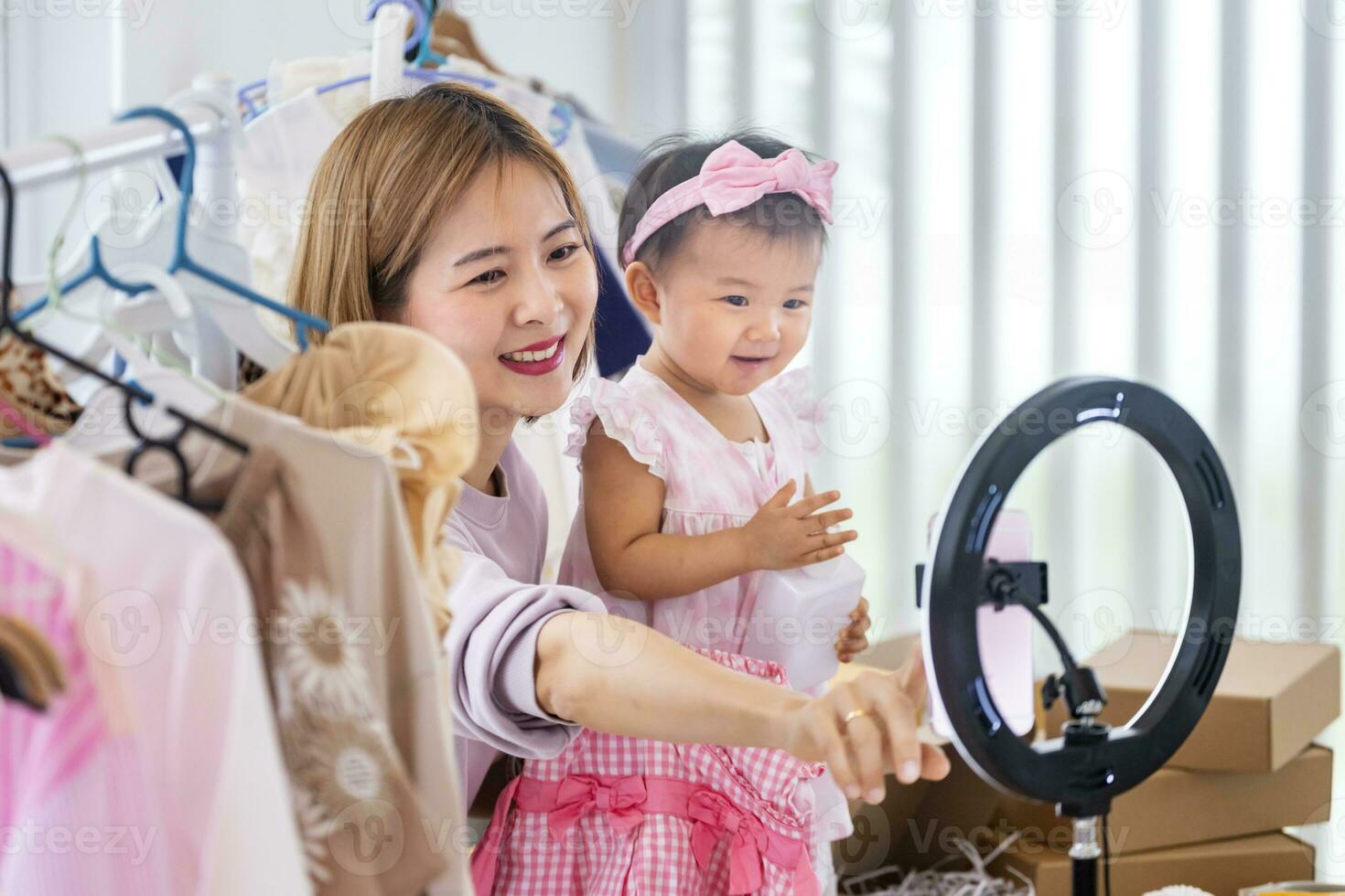 jung asiatisch Influencer Mutter ist Verkauf das Kinder Kleider Leben online Streaming beim Zuhause während erziehen ihr Kleinkind wie Hilfe foto