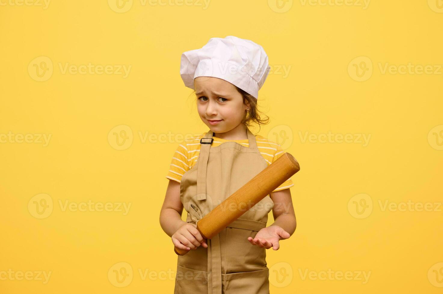 Studio Porträt von erstaunt wenig Mädchen, Koch Gebäck, im Weiß Deckel und Beige Schürze, hoding rollen Stift, isoliert auf Gelb foto