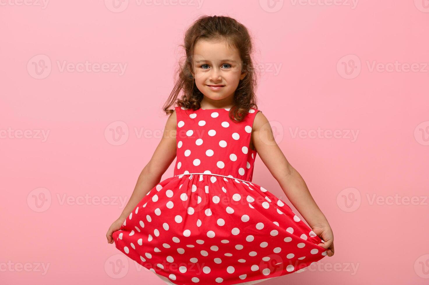Porträt von süß charmant Mädchen im ein Rosa Kleid mit Weiß Polka Punkte posieren onpink Hintergrund süß lächelnd und halten das Unterseite von das Kleid mit ihr Hände. schön Kind Porträt zum Werbung foto