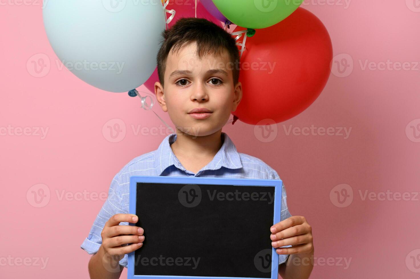 gut aussehend Junge mit leer Tafel im seine Hand Stehen gegen mehrfarbig Ballons auf Rosa Hintergrund mit Kopieren Raum foto