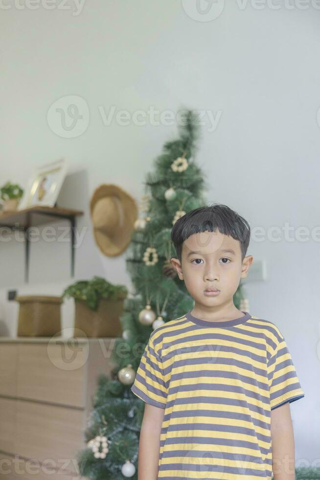 Nahansicht asiatisch Junge mit schwarz Haar tragen ein gelbgrau gestreift T-Shirt suchen beim das Kamera im das Hintergrund von Weihnachten Bäume und hölzern Möbel. Kind macht komisch Gesichter im seine Haus glücklich. foto