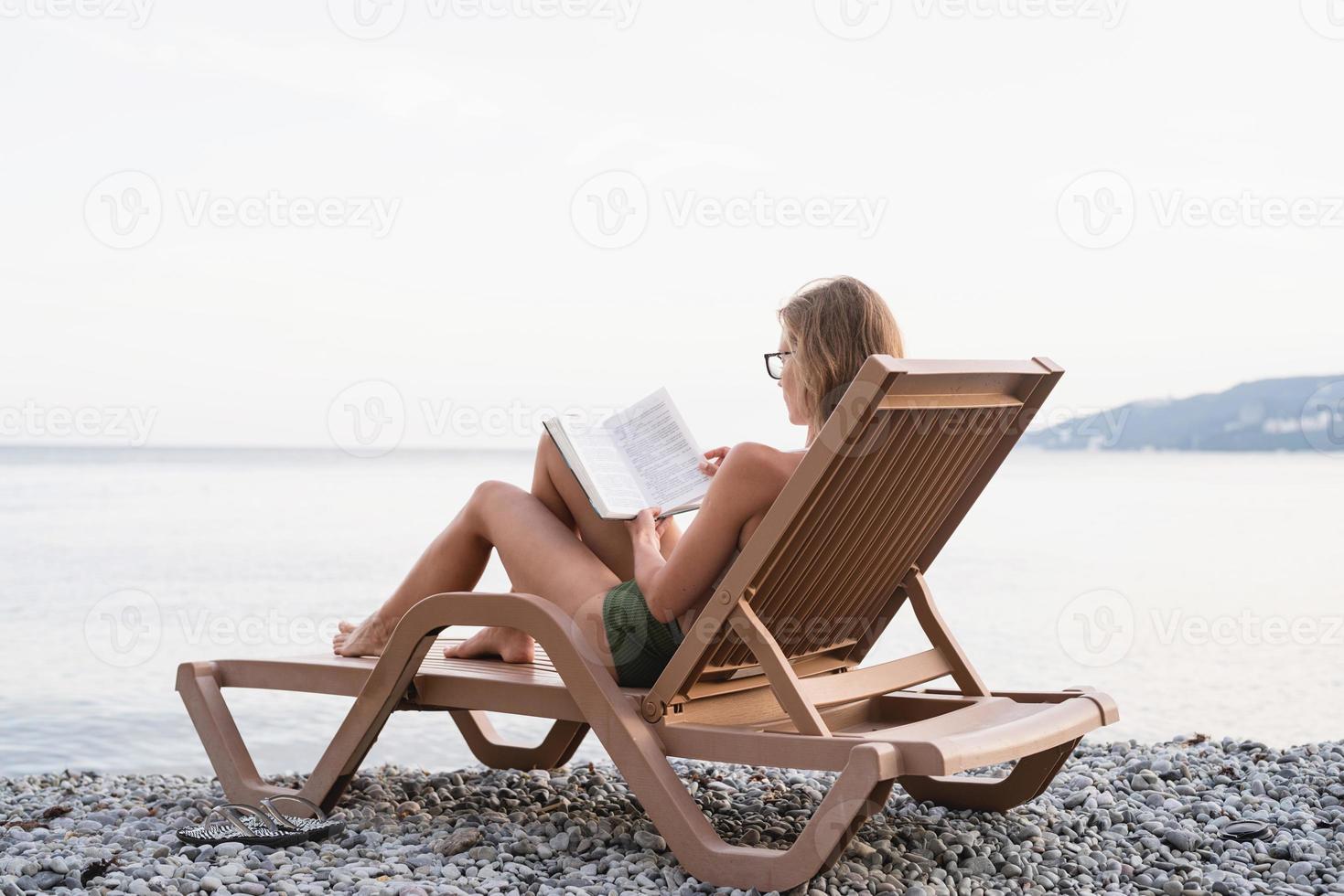 Die schöne junge Frau sitzt auf der Sonnenliege und liest ein Buch foto