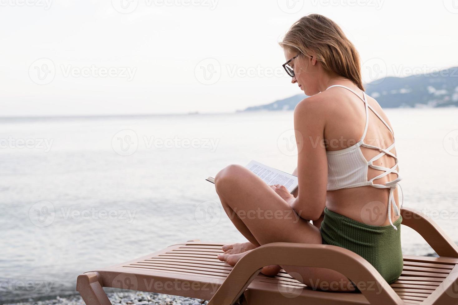 Die schöne junge Frau sitzt auf der Sonnenliege und liest ein Buch foto