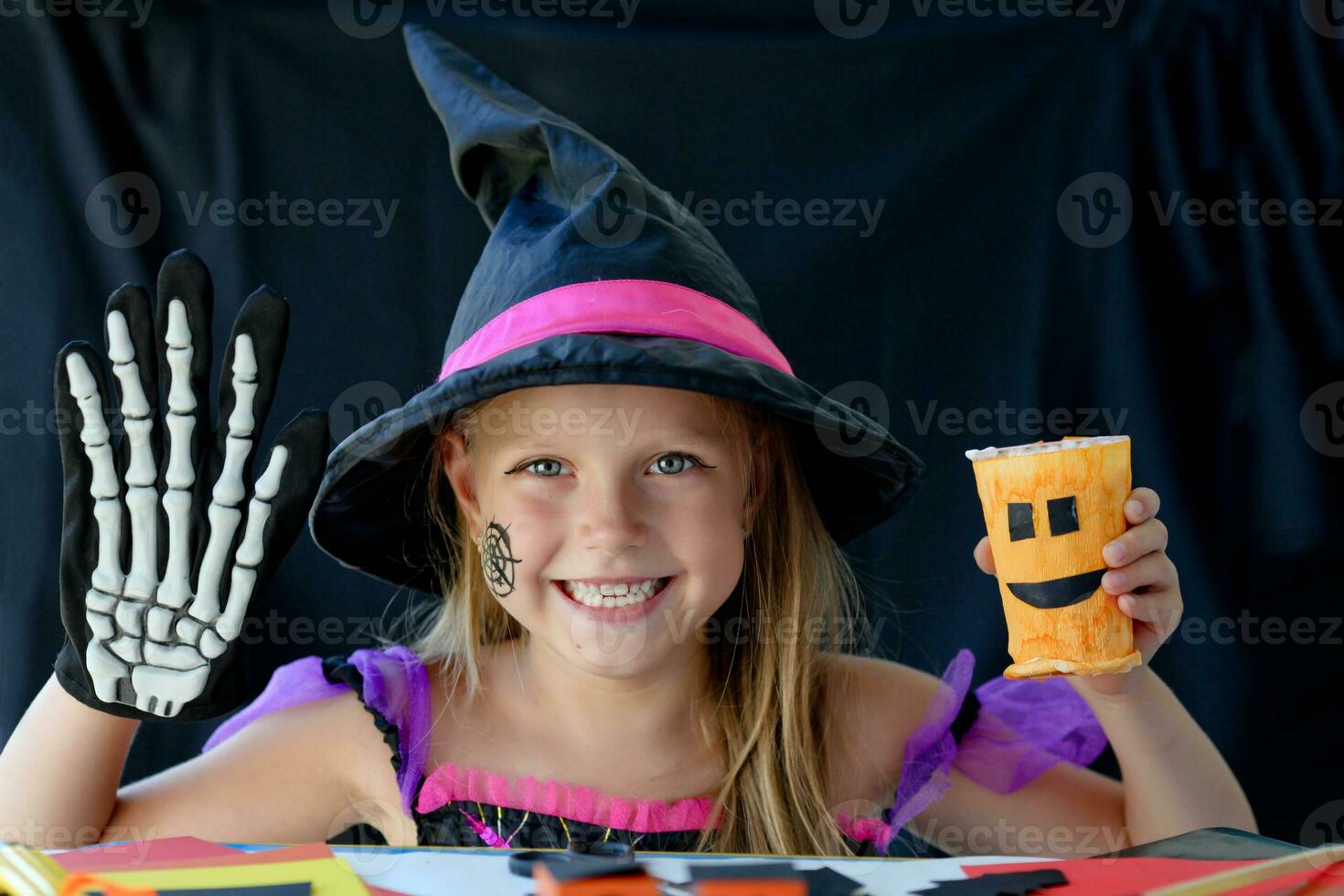 ein wenig Mädchen im ein Hexe Kostüm sieht aus beim das Kamera und lacht, halten ein Glas Kürbis und ein Skelett Handschuh im ihr Hände auf schwarz Hintergrund. foto