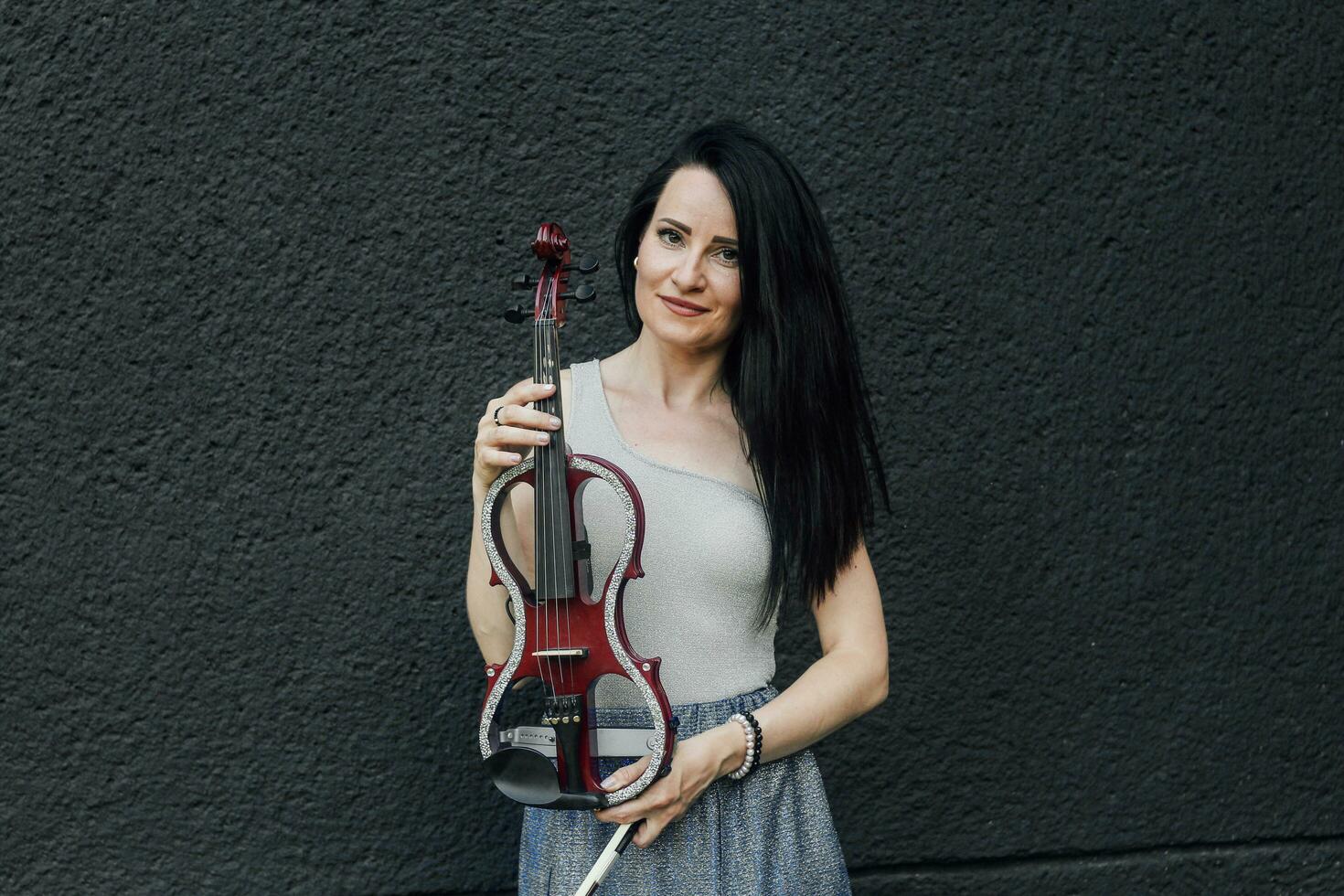 ein Frau Künstler mit dunkel Haar im ein Kleid hält ein hölzern Konzert elektrisch Violine im ihr Hände foto