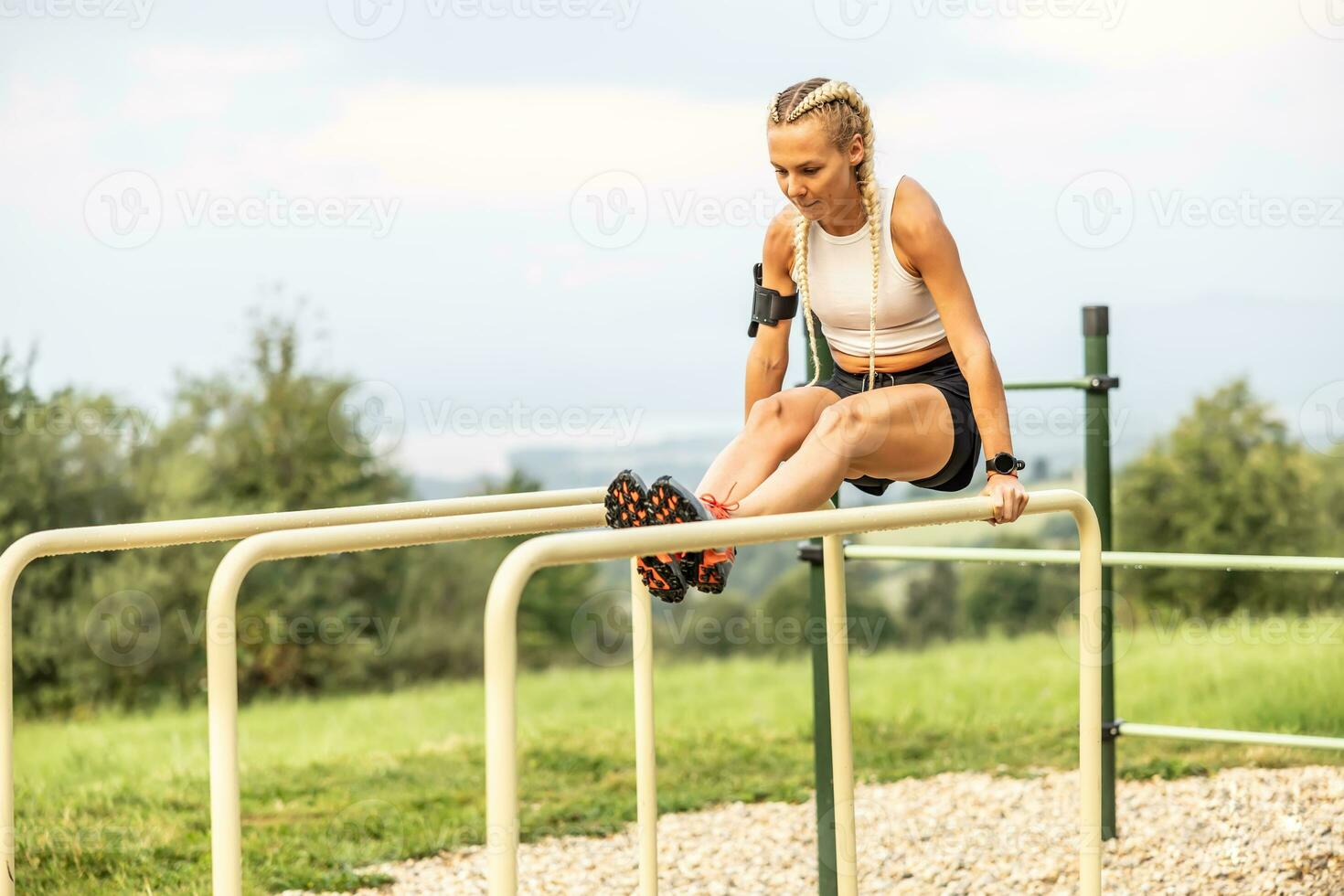 sportlich jung weiblich Arbeiten aus Gymnastik im ein draussen Fitnessstudio mit parallel Riegel. schön blond Mädchen mit Zöpfe funktioniert aus draußen tun Liegestütze auf ein erhöht Bar foto