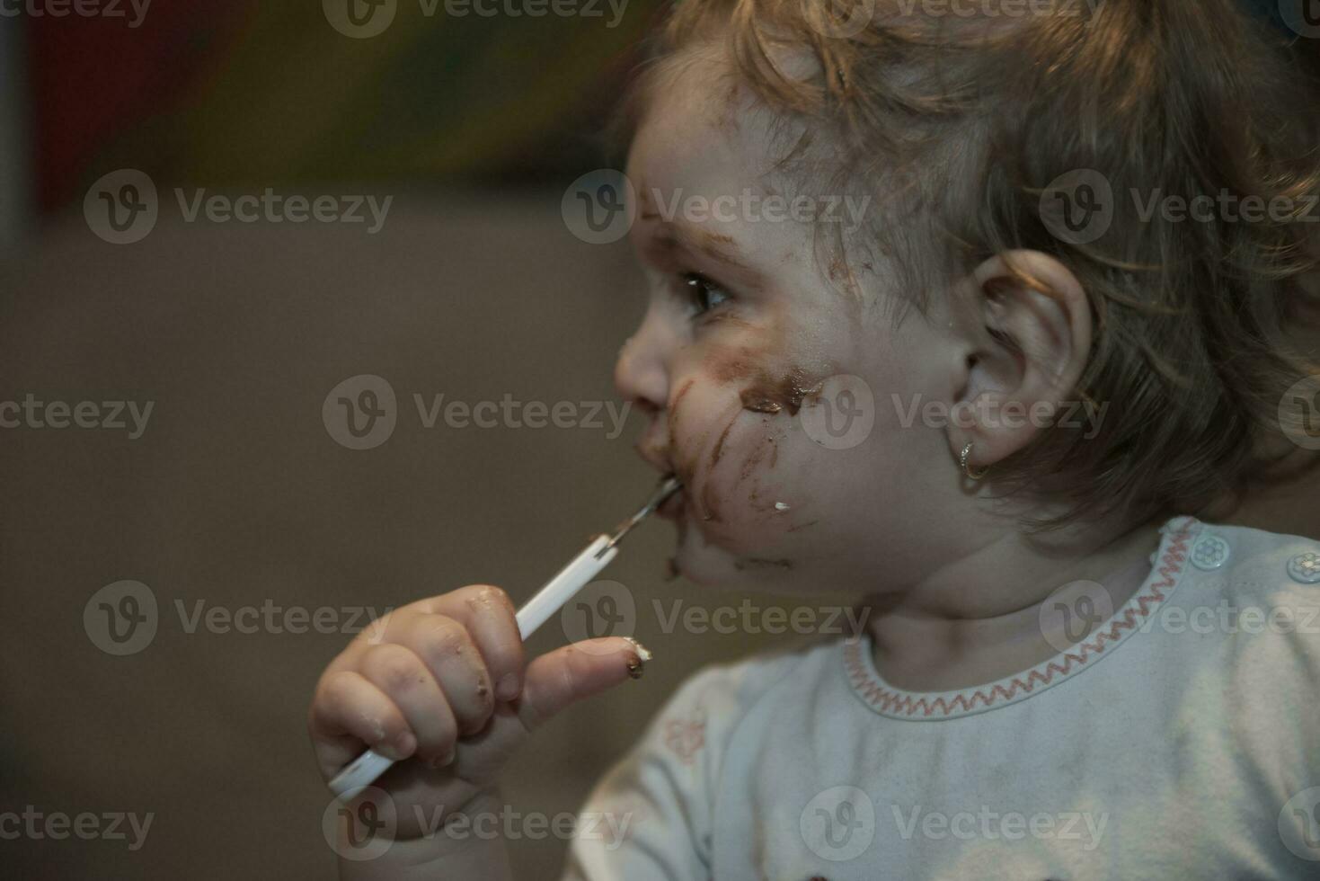 Baby Mädchen Essen ihr Schokolade Wüste mit ein Löffel und Herstellung ein Chaos foto