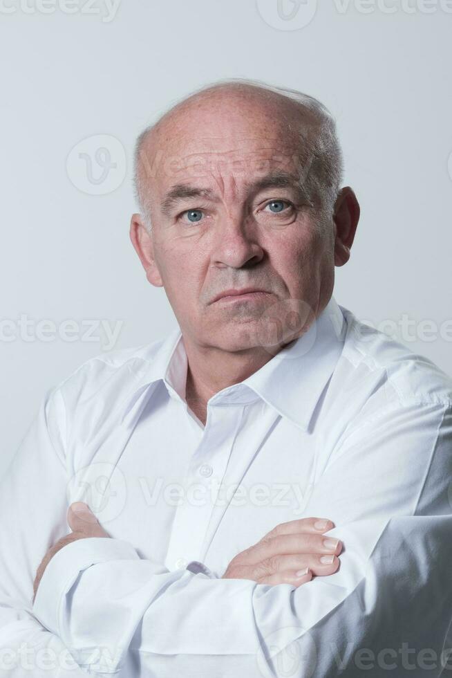 zuversichtlich Senior Mann im Weiß Hemd Kreuzung Hände auf Truhe und suchen beim Kamera während Stehen gegen grau Hintergrund. selbst zuversichtlich Senior isoliert Weiß Studio schießen. foto