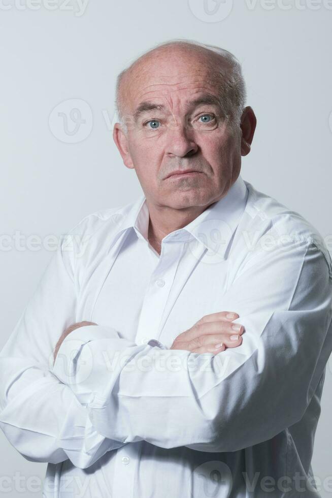 zuversichtlich Senior Mann im Weiß Hemd Kreuzung Hände auf Truhe und suchen beim Kamera während Stehen gegen grau Hintergrund. selbst zuversichtlich Senior isoliert Weiß Studio schießen. foto