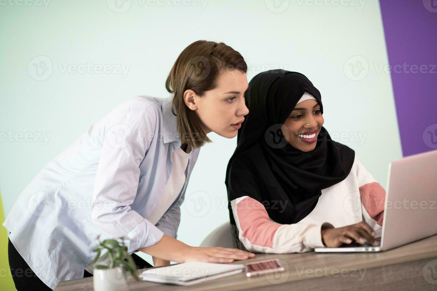 afrikanisch amerikanisch Frau mit ein Hijab und ein europäisch Frau mit ein Smartphone und Laptop im ihr Zuhause Büro foto