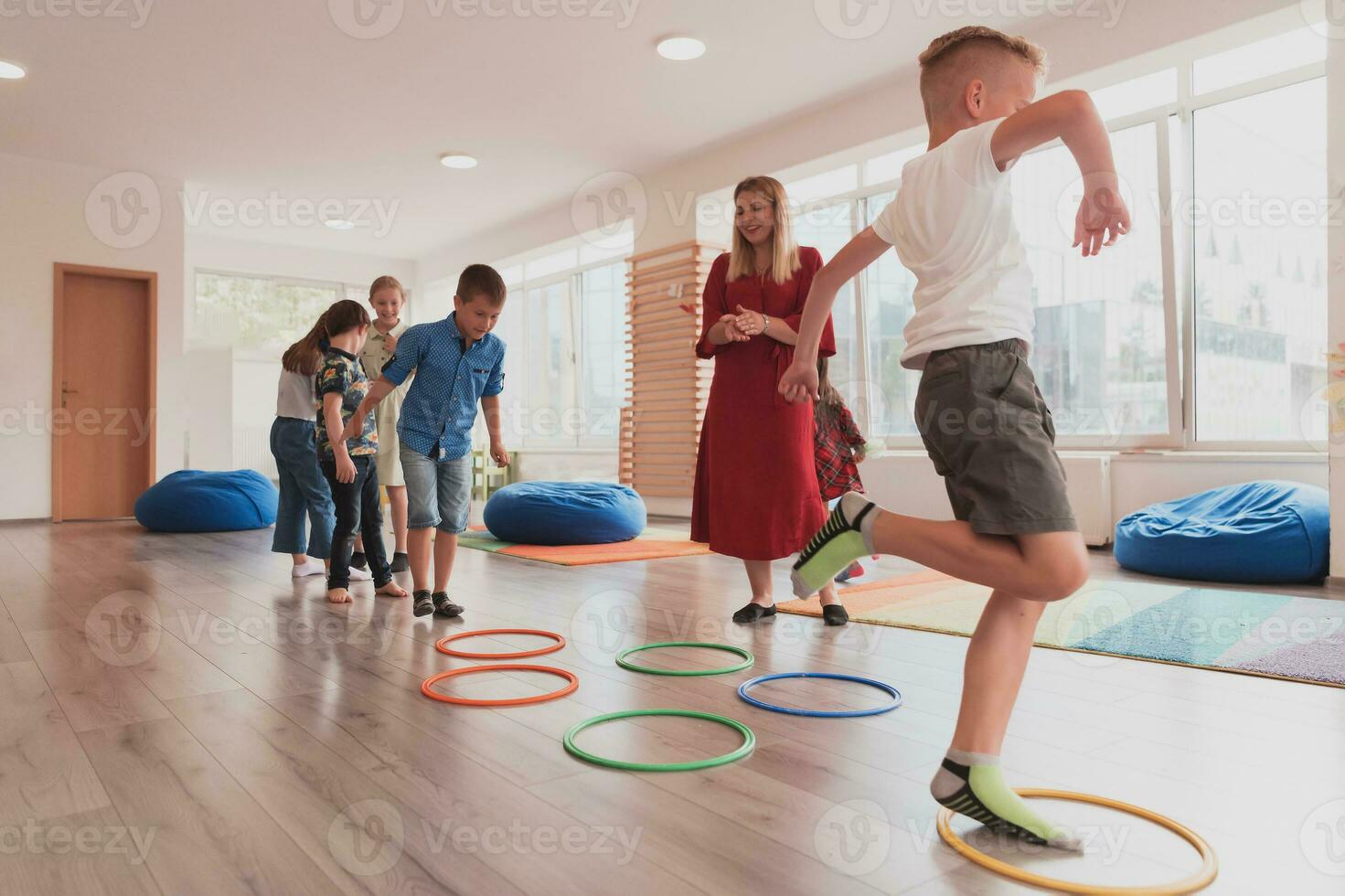 klein Kindergarten Schule Kinder mit weiblich Lehrer auf Fußboden drinnen im Klassenzimmer, tun Übung. Springen Über Hula Band Kreise Spur auf das Boden. foto