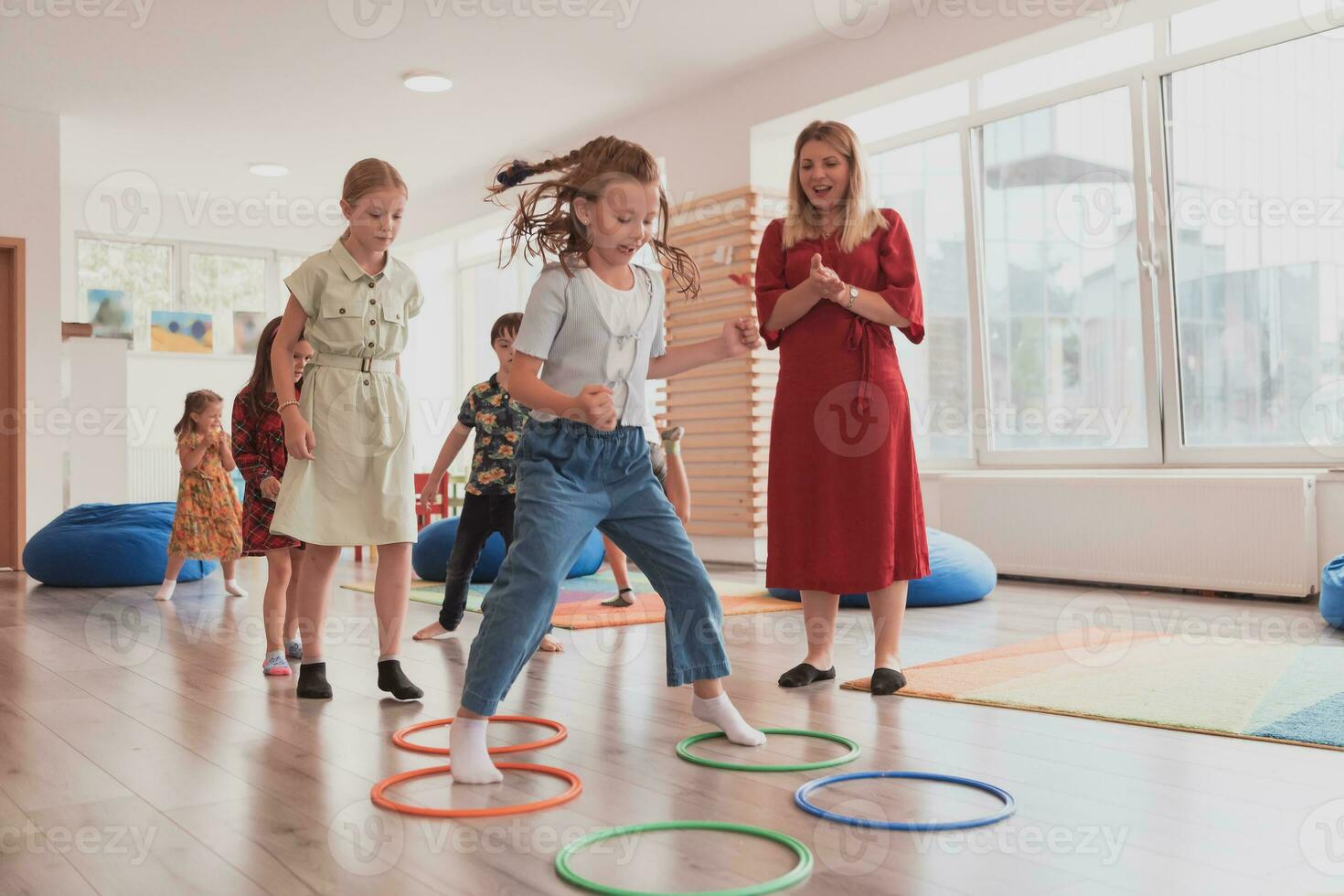 klein Kindergarten Schule Kinder mit weiblich Lehrer auf Fußboden drinnen im Klassenzimmer, tun Übung. Springen Über Hula Band Kreise Spur auf das Boden. foto