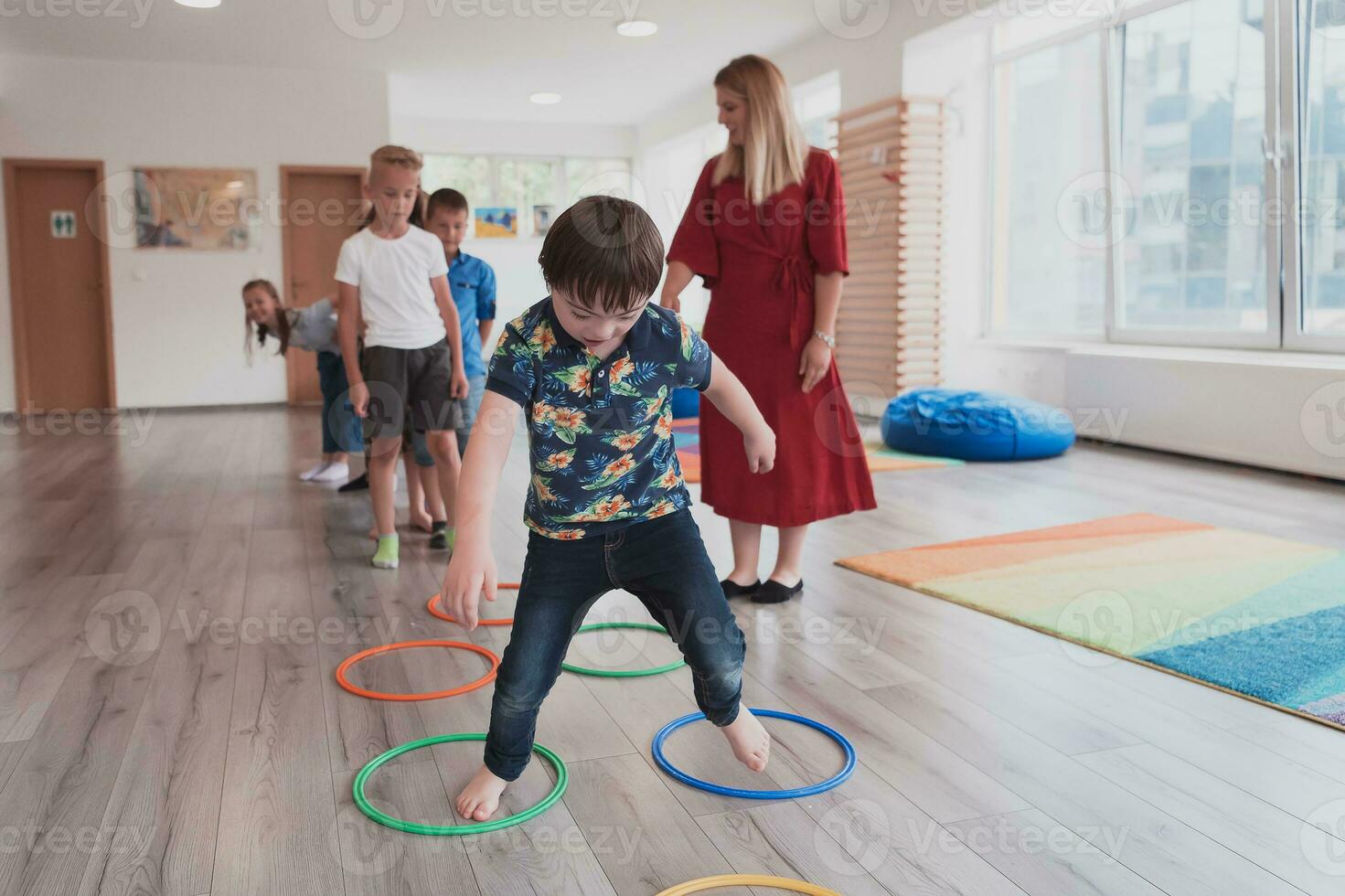 klein Kindergarten Schule Kinder mit weiblich Lehrer auf Fußboden drinnen im Klassenzimmer, tun Übung. Springen Über Hula Band Kreise Spur auf das Boden. foto