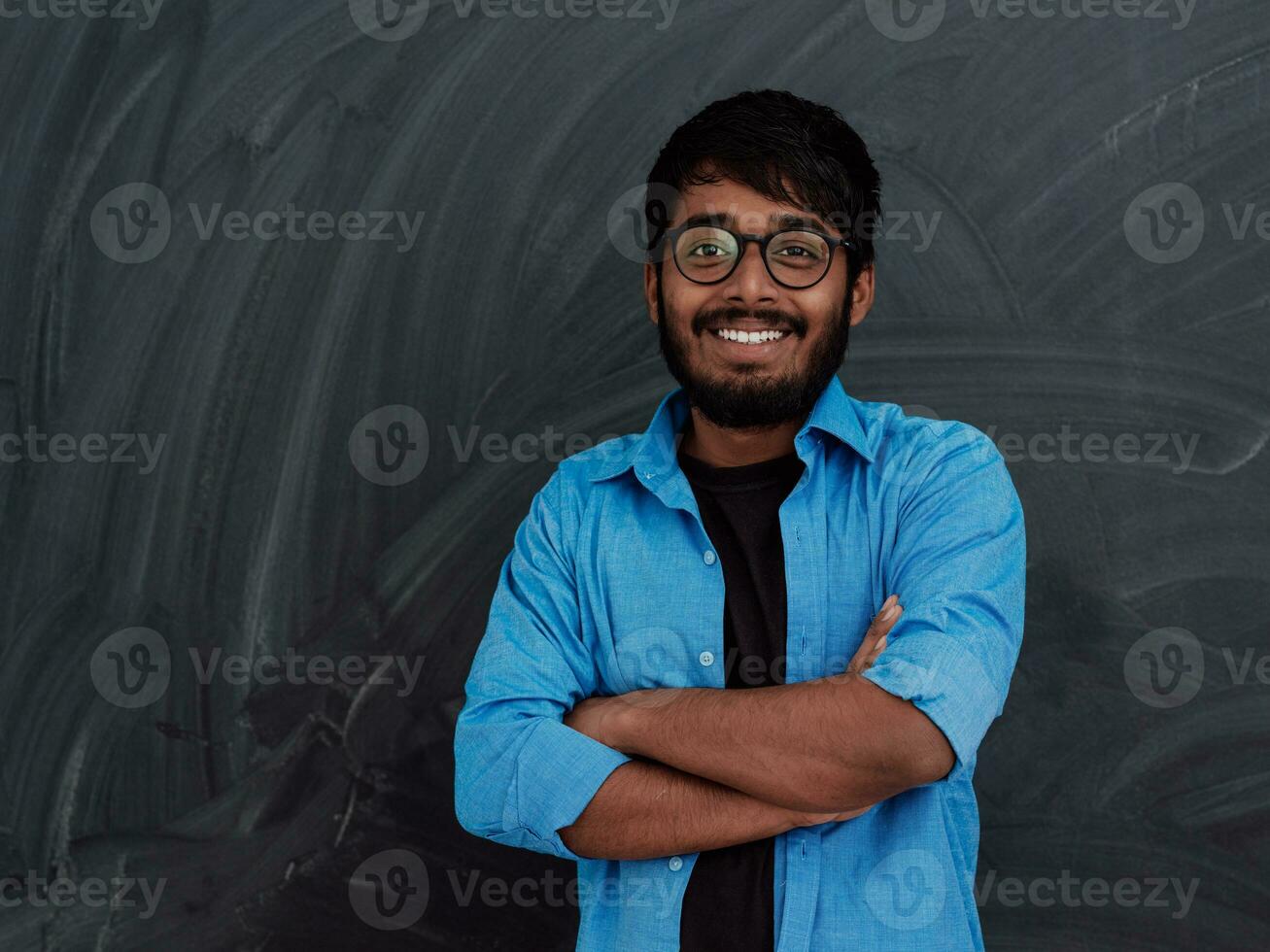 ein jung indisch Schüler im ein Blau Hemd mit Brille posieren mit seine Waffen gekreuzt im Vorderseite von das Schule Tafel foto